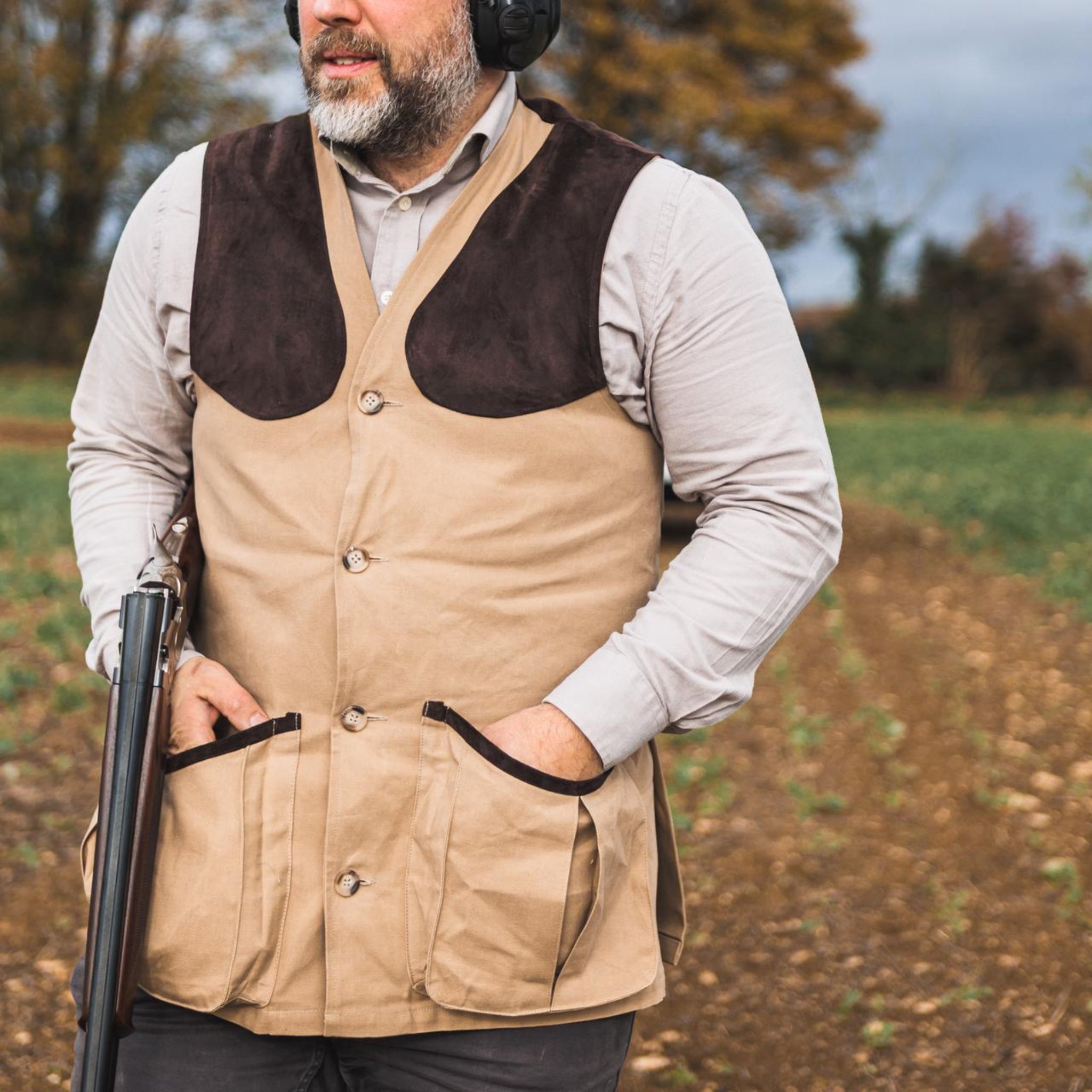 Man wearing a shooting vest and ear defenders, holding a twelvebore shortgun in an outdoor setting.