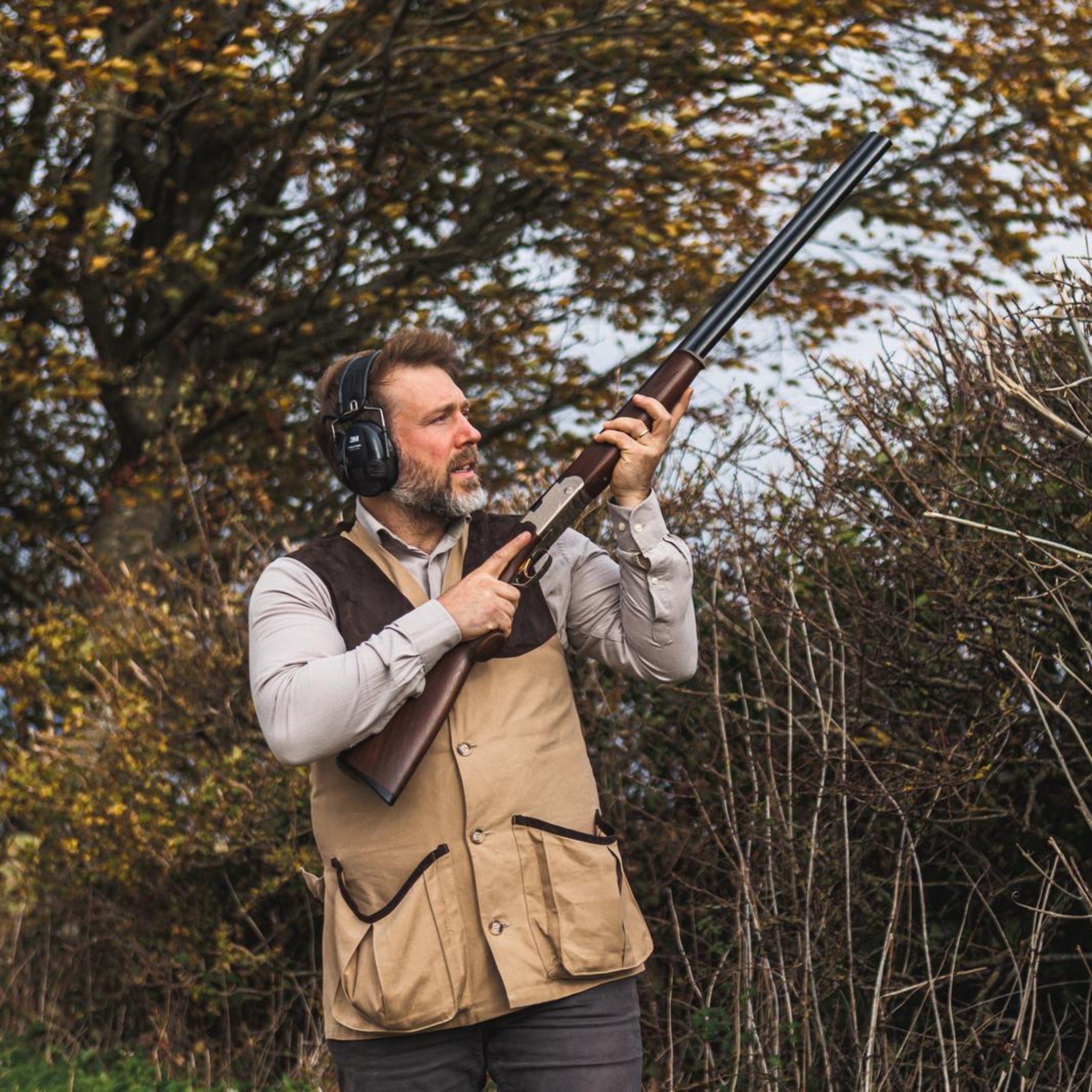 Man holding a shotgun in a natural outdoor setting with trees and shrubs.